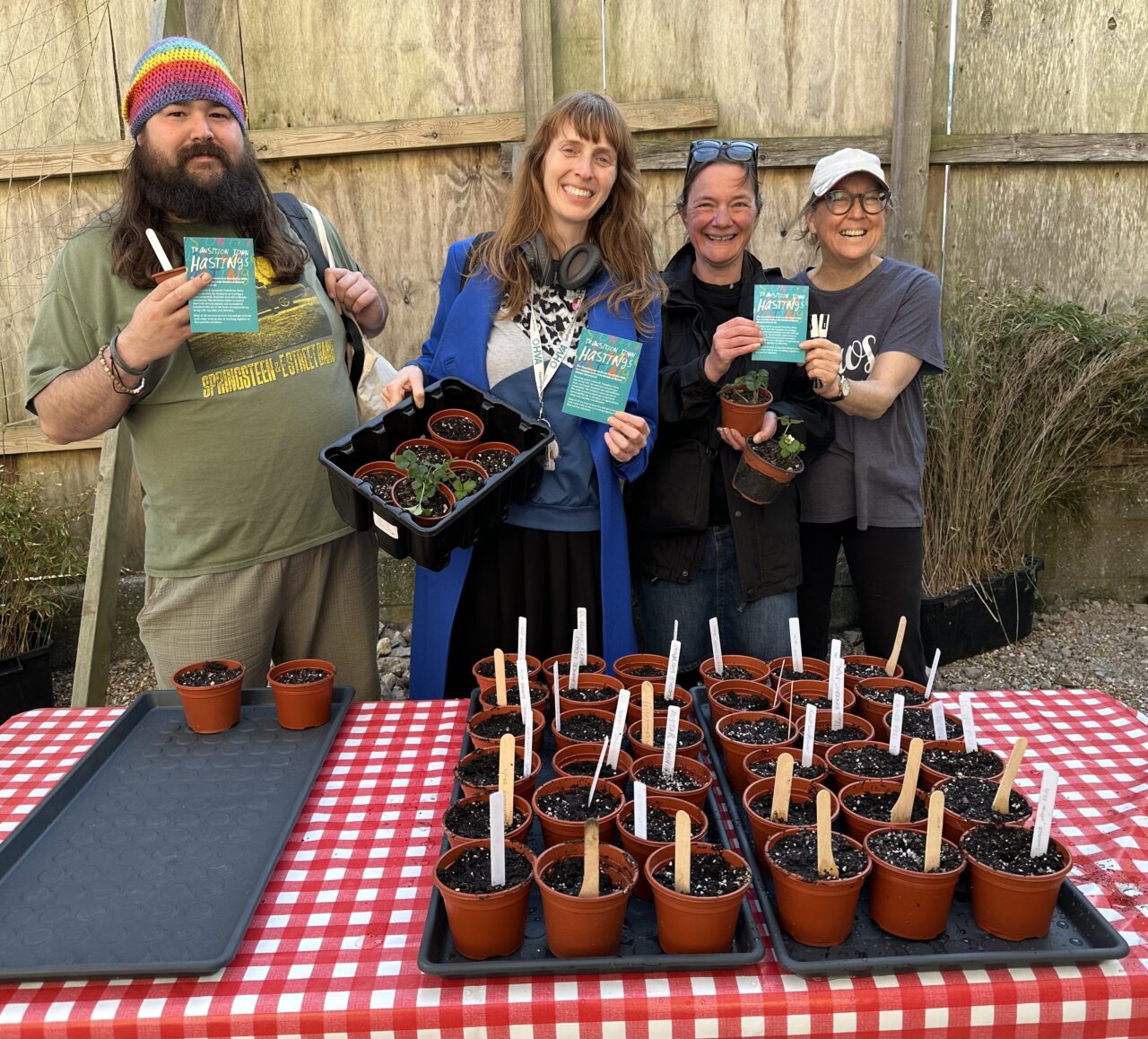 Jane from HVA standing with 3 community members, all holding up packets of seeds and standing in front of a table with lots of potted seeds, all smiling at the camera