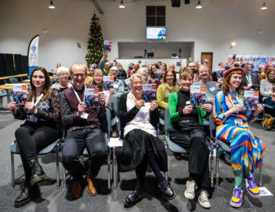 A group of the HVA team sitting in a row of 5 seats, behind them there are a lot of other people sitting in seats, all smiling and holding up leaflets, towards the camera