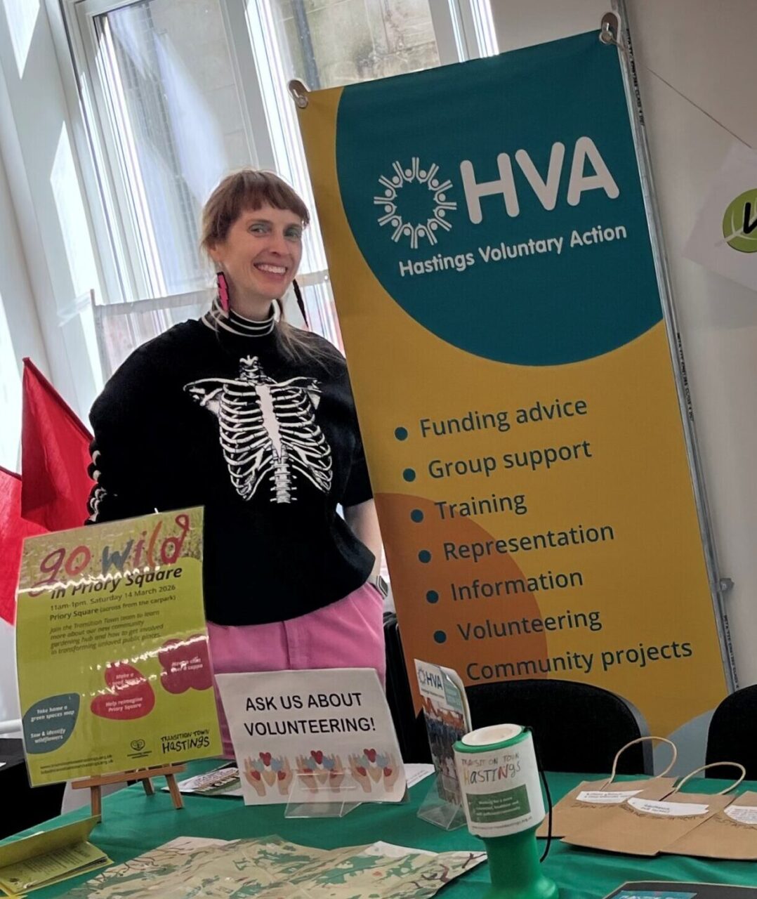 Jane Caley standing at a table, advertising HVA and volunteering. Jane is wearing a black and white top with pink trousers
