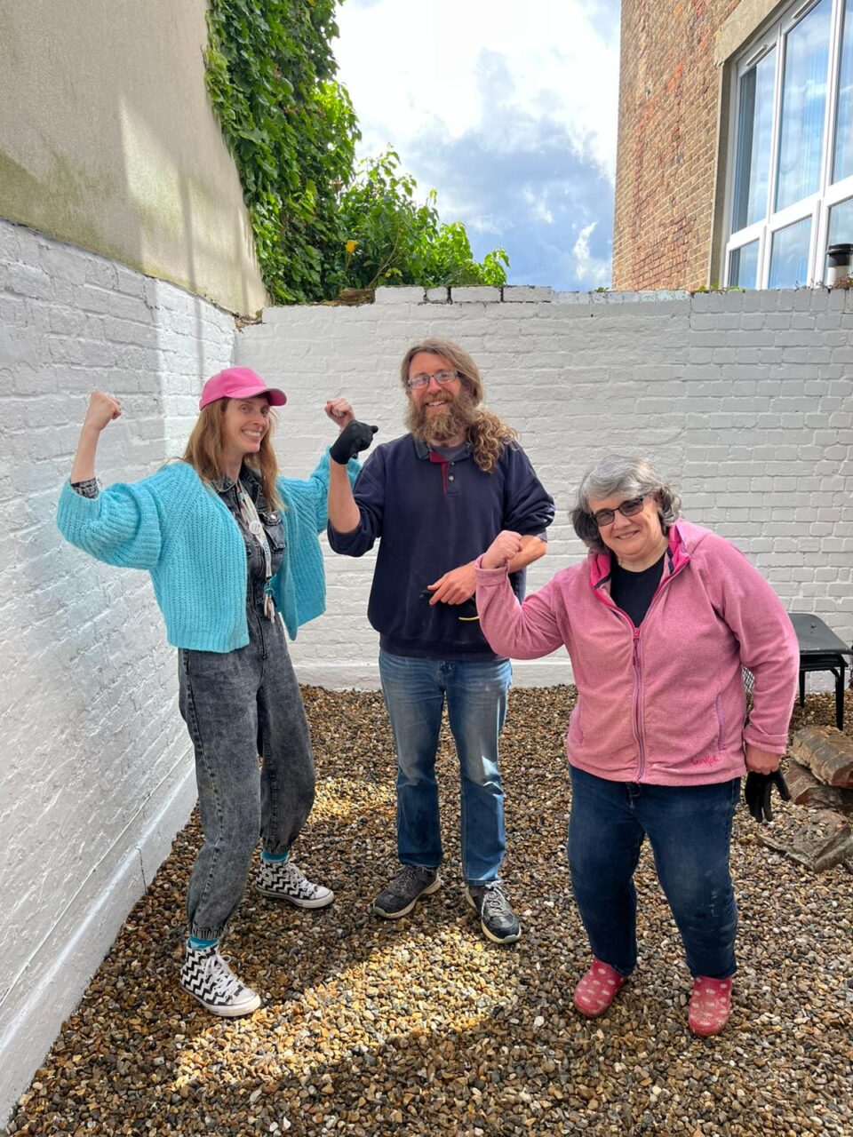 3 people standing in a courtyard garden space showing off their muscles after moving lots of gravel